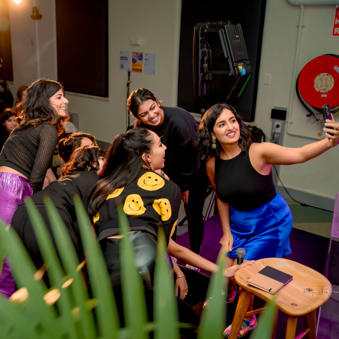 A group of women performers gather onstage, smiling and posing for a selfie after a show.”